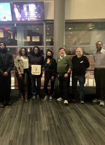 A diverse group of seven individuals, four men and three women, take a group photo inside a room with large windows. An African American woman, third from left, holds a SAG-AFTRA canvas bag. Above head are two television screens.