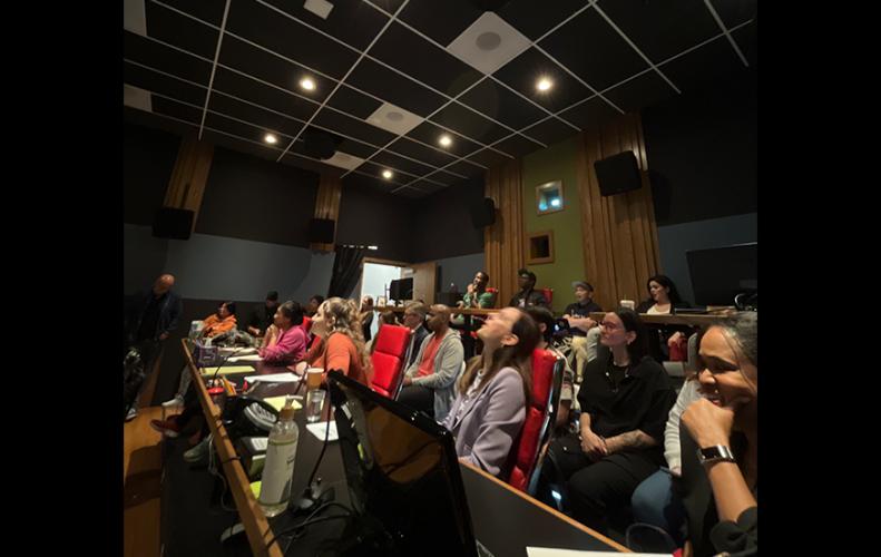 A diverse crowd seated at long tables within a small auditorium. They are arranged in a few rows and look offscreen to the left. Also visible are speakers, some computer screens and scripts.