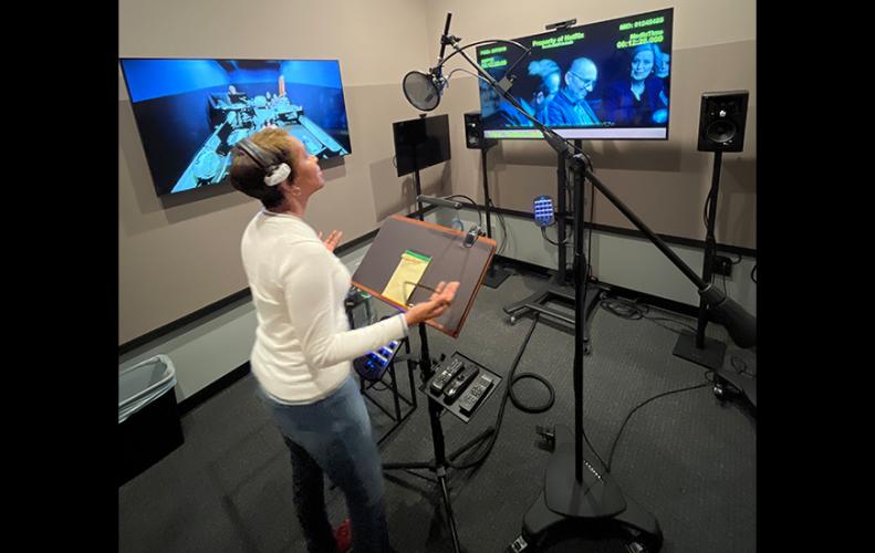 Monteith, a woman of color, is in a vocal recording studio with sound recording equipment. She is dressed casually and looks at a flatscreen that displays a scene from a movie, gesturing with her hands as she recites her lines.