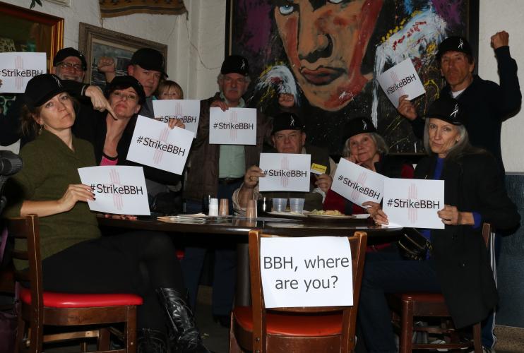 Members each hold a #StrikeBBH paper while seated around a brown table and one empty chair that signifies BBH