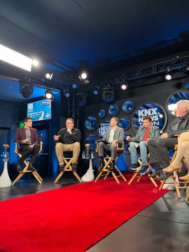 Crabtree-Ireland, seated second from left, speaks. He wears a black jacket with SAG-AFTRA’s logo and khaki pants. Other panelists are visible. The studio has bright lights, KNX’s logo, and a red carpet on the floor.