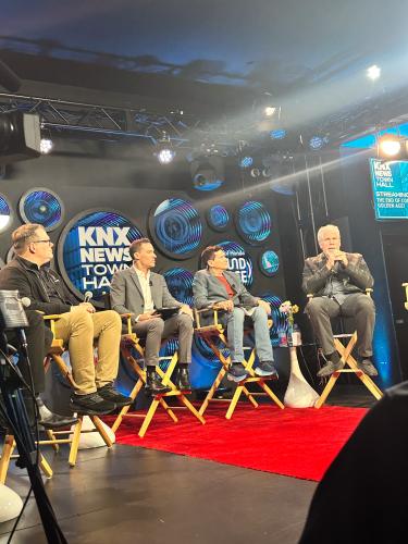 Perlman, seated on the far right, speaks. He wears a black leather jacket and dark jeans. Other panelists are visible. There is a glare from a studio light; KNX’s logo on the back wall and a red carpet on the floor.