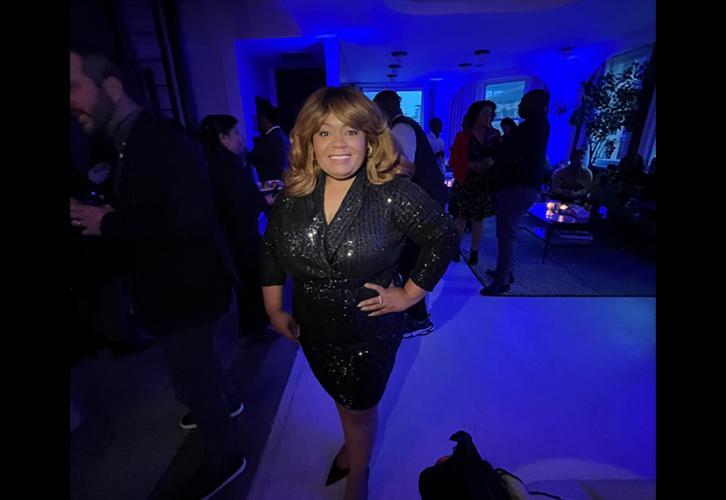 Underdue (African American) stands in a dimly lit room. She wears a black dress and her hands are on her hips. She smiles at the camera.