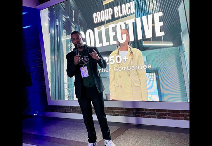 Trimble (African American) stands in front of a screen with a Group Black Collective title card. He wears a coat over a green hoodie and jeans, and holds a microphone.