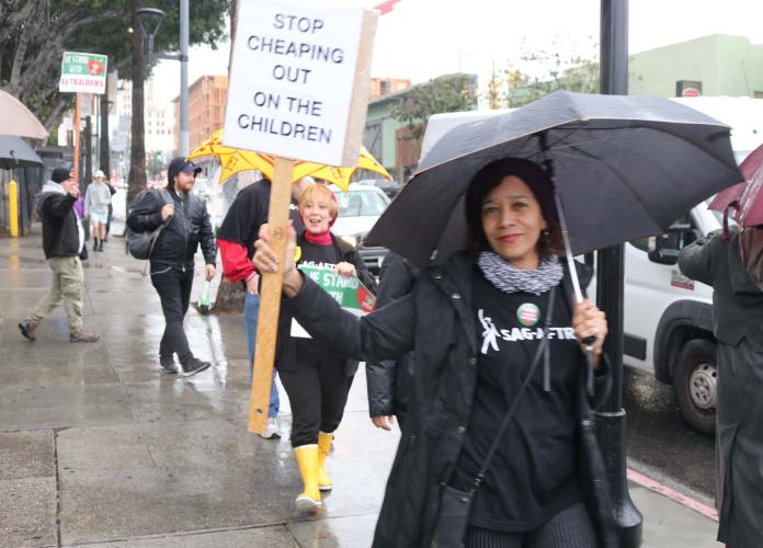 SAG-AFTRA member wearing a t-shirt and holding an umbrella in her left and a sign in her right that reads "STOP CHEAPING OUT ON THE CHILDREN"