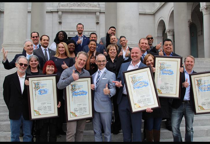 A large group of men and women display certificates of recognition and give the camera a big thumbs up. 