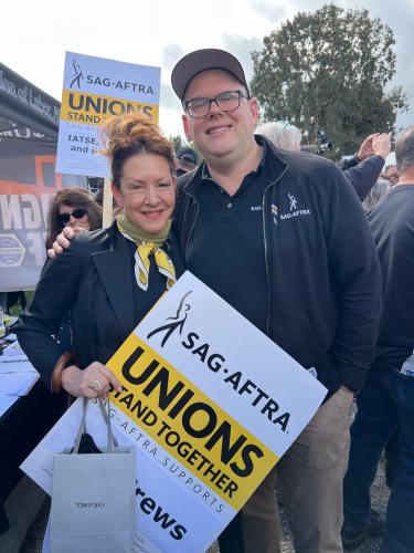 Fisher, left, and Crabtree-Ireland stand closely for a photo. Both wear black with Fischer in an all-black outfit with a yellow bandana around her neck. Fisher holds a SAG-AFTRA picket sign and a small gray shopping bag.