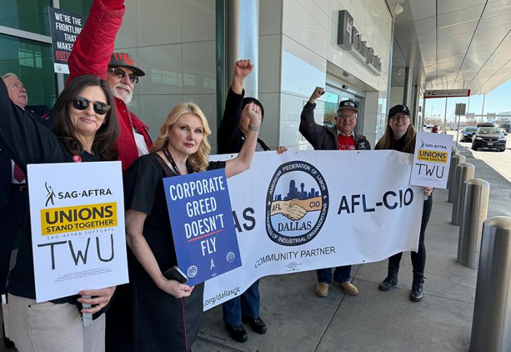 Un grupo de personas afuera de un edificio anodino, posiblemente un aeropuerto. Cuatro personas, en el extremo derecho, sostienen una pancarta de la AFL-CIO de Texas; otros sostienen carteles SAG-AFTRA o piquetes hechos a mano. Cada uno levanta un puño solidario.