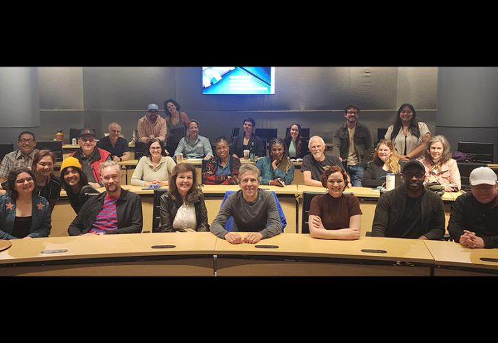 A group photo of people of different ethnicities in a boardroom. The group is arranged in three rows and seated behind long tables. All wear casual clothing.