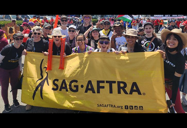 A large group of  people of different ethnicities pose with a yellow SAG-AFTRA banner. Many wear SAG-AFTRA or "SAG-AFTRA Strong" T-shirts.