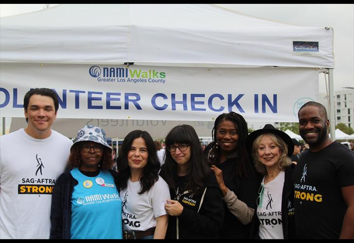 Seven people of different ethnicities stand outside of a tent. Aside from a Black woman, second from left, who wears a sky blue NAMIWalks t-shirt, all wear black or white SAG-AFTRA T-shirts.  The tent banner reads “Volunteer Check In” in all caps.
