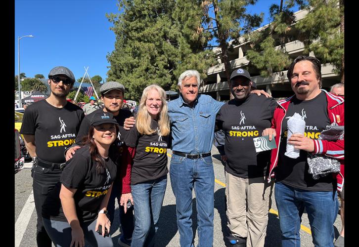 An outdoor group photo of men and women of different ethnicities wearing black "SAG-AFTRA Strong" shirts. Jay Leno, center, is dressed in a long sleeve denim shirt and jeans.