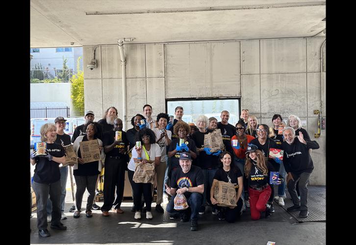 A large group photo people  from different ethnicities  take a photo inside a warehouse building. All hold a non-perishable food item; some hold paper grocery bags.