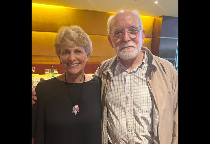 Karas and Stanley stand close together. Karas wears a black top and necklace; Stanley wears a brown jacket, a graph-patterned shirt and khakis. The lighting is warm.