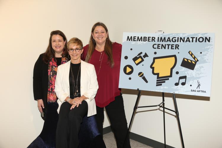 Carteris sitting cross legged on a chair with Damon over Carteris' right shoulder and Manheim over Carteris' left shoulder with a "Member Imagination Center" poster to Manheim's left