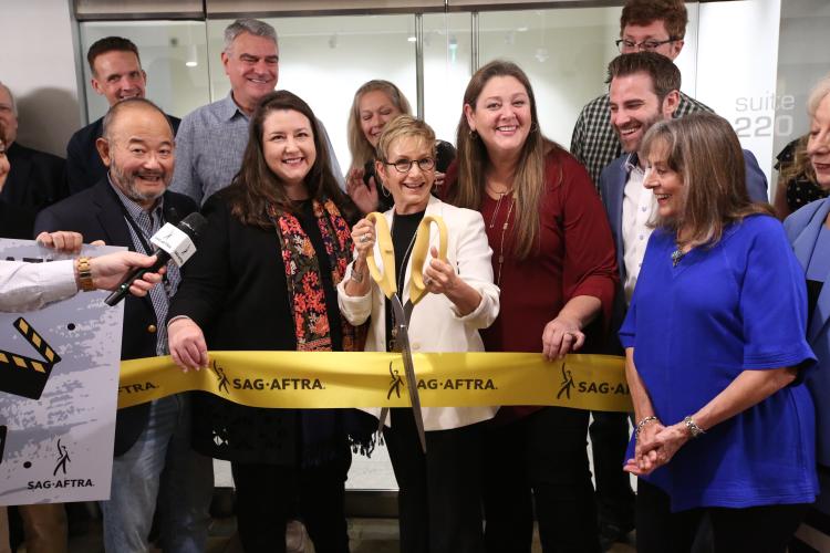 Carteris holding big scissors to cut yellow SAG-AFTRA ribbon with members all around.