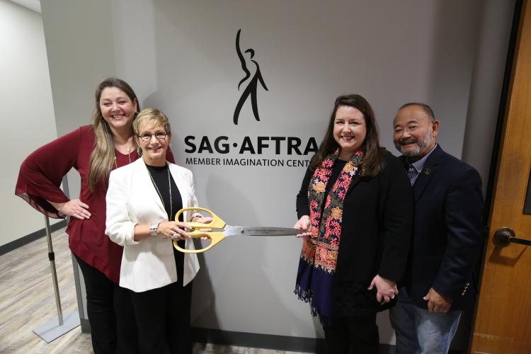 Manheim in Maroon standing behind Carteris in black with a white blazer holding the handle of large scissors. Damon holding the opposite end of scissors wearing black and a colorful scarf with Kusatsu standing behind her. "SAG-AFTRA Imagination Center"
