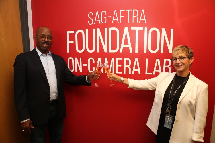 Vance in a black suit with a light blue button up holding a champagne flute cheersing Carteris in a white blazer and black top standing in front of a red wall with the "SAG-AFTRA Foundation" logo in white and "On-Camera Lab" in white below the logo.