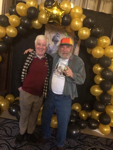 O’Neill and Samuels stand under a balloon arch. O’Neill’s outfit is business casual; Samuels is dressed casually with a colorful helicopter cap. He points to the camera.