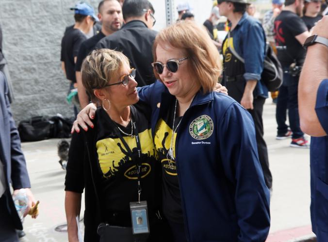 Carteris in a black #SOSSBS t-shirt wraps left arm around Durazo's shoulder and Durazo in black #SOSSBS t-shirt and navy blue jacket holds right arm around Carteris's shoulder
