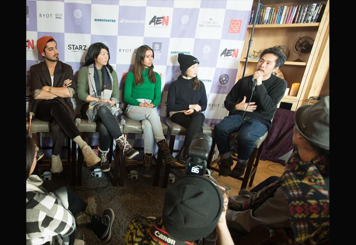 A group of men and women seated in chairs listens to an individual, male, speak. 