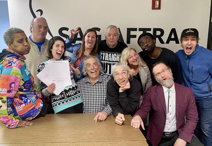 People gather around a table and make funny faces for the camera. There is a wall with the SAG-AFTRA logo behind them.