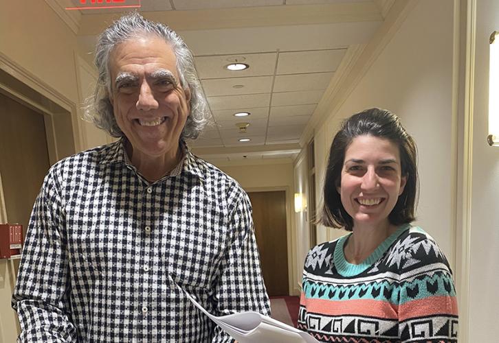 Rossi, in a black-and-white dress shirt, and Feeney, in a colorful sweater, both smile while standing in a hallway.