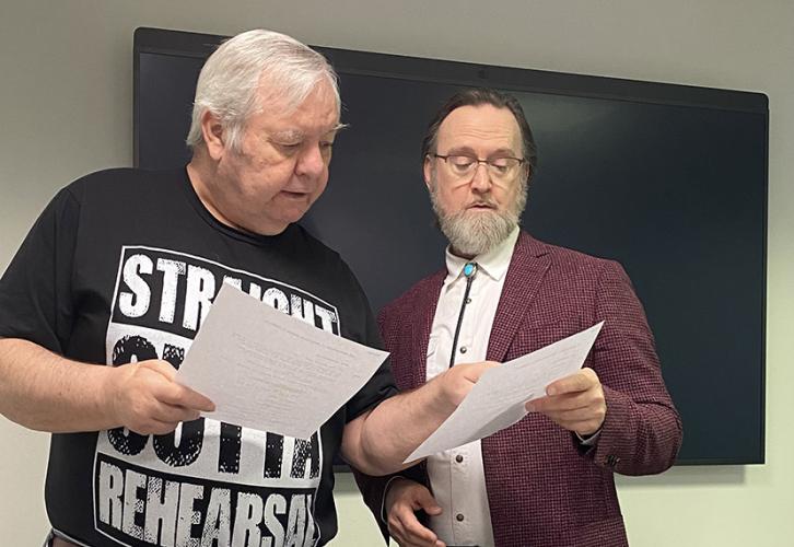 Viens, in a "Straight Outta Rehearsal" T-shirt, points to something on Rudolph's script. Rudolph is wearing a coat, dress shirt and bolo tie.