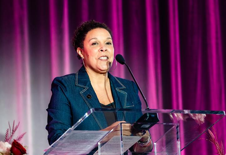 Powell stands at a podium. She wears a jean blazer and a black shirt underneath. She is captured mid-speech and staring out into the audience. Behind her is a dark pink curtain.