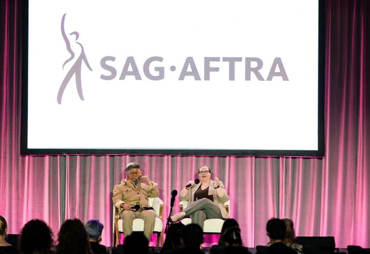 Stivale and Johnson are seated in chairs on stage. Johnson is caught mid-sentence; she holds up a microphone with her right hand and gestures with her left. Stivale looks on, his body positioned to face Johnson. Behind them is a dark pink curtain; above them is a projector screen bearing the SAG-AFTRA logo. Some audience members are visible in the foreground.
