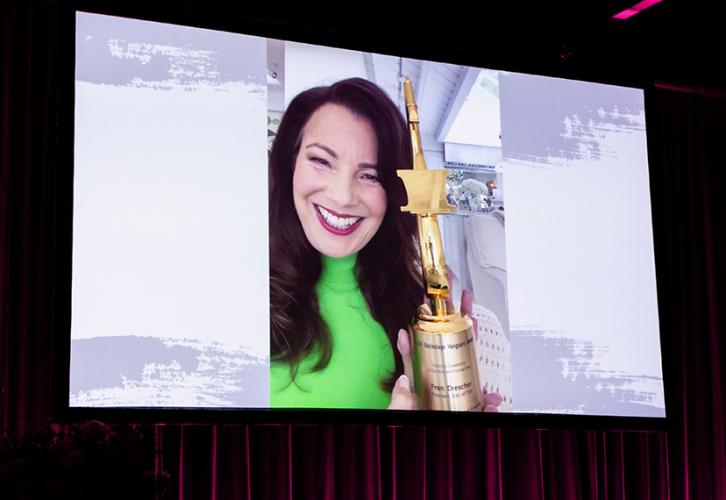 A video clip of Drescher holding a large gold statuette with text on its base. She wears a lime green turtleneck shirt. Bordering the image is a gray background with dark-colored brush strokes.