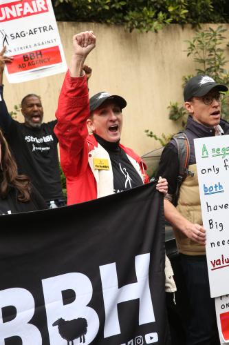 Austin in a red rain coat, black t-shirt and black Ads Go Union hat holds her right fist in the sky while chanting and holding a #StrikeBBH banner in her left hand. 