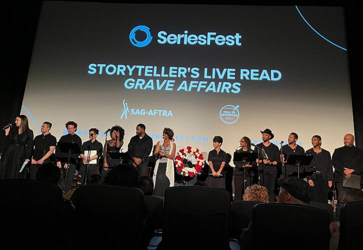 Trece personas suben al escenario, a cada lado de una corona de flores. Detrás de ellos, hay una pantalla grande con las palabras "SeriesFest, Storyteller's Live Read 'Grave Affairs'".