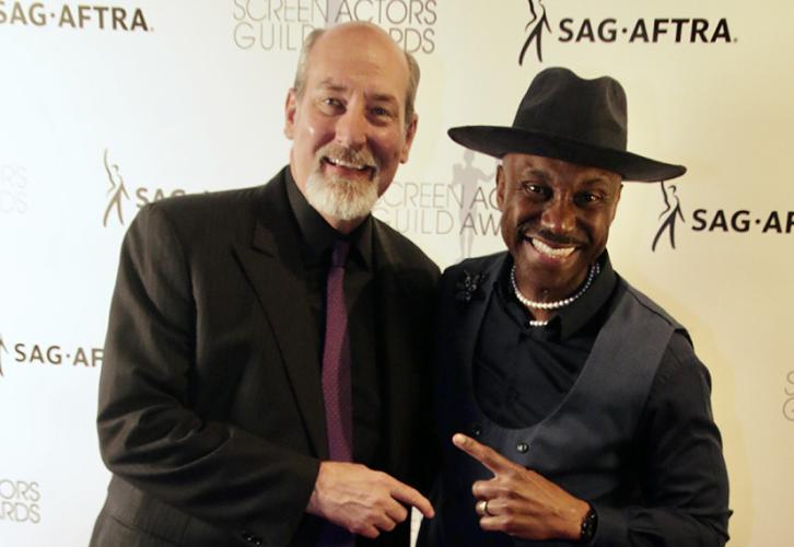 Two men smile and point to each other. One wears a black hat.