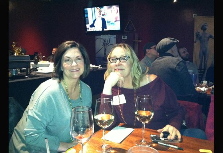 Two women at a table with drinks smile at the photographer. 