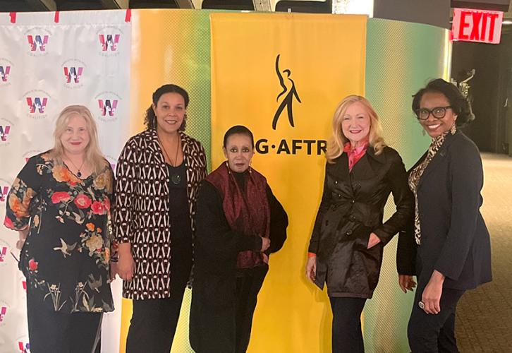 A group of five individuals are in front of a white step-and-repeat backdrop with the Women in Arts & Media logo on the left, and a yellow standing SAG-AFTRA banner on the right. 