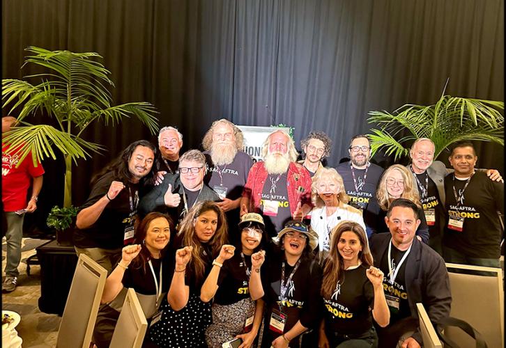 A diverse group takes a photo on stage in a large conference hall. Those in the first row are crouched and many raise a fist in solidarity. A spotlight or projector screen is centered on those in the back row. The group is framed by indoor palm trees. 
