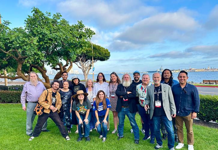 A large diverse group of individuals take a group photo outside. Two women in front crouch slightly. Also visible is grass underfoot, a tree, far left, and a cloudy sky.