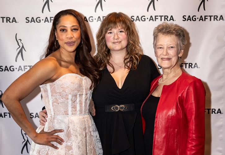 Una mujer con un vestido blanco, una mujer con un vestido negro y una mujer con un abrigo rojo posan frente a un telón de fondo con logotipos de SAG-AFTRA.