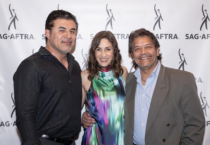 Una mujer con un vestido colorido se encuentra entre dos hombres frente a un fondo con logotipos de SAG-AFTRA.