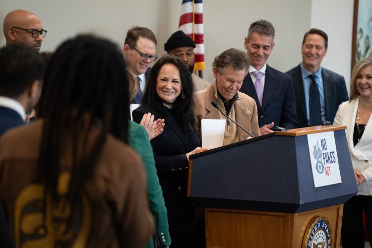 Mary Travis and Randy Travis standing behind a podium adorned with a NO FAKES Act sign