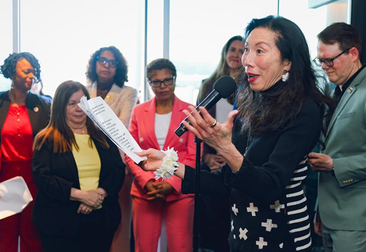 Jodi Long is in front of a microphone. She is captured mid-speech with a set of papers in her right hand. She gesticulates with her left hand. Bystanders look on. 