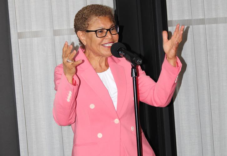 Karen Bass speaks at a microphone. They are wearing a pink pantsuit. As they speak, they gesticulate with their hands and smile.