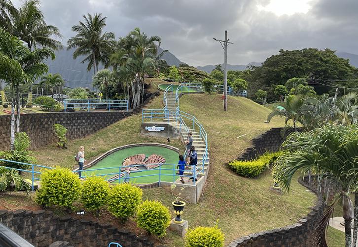 The last hole of the mini golf course is set along a hillside. There are concrete stairs with a bright blue railing along the hillside leading to and away from the green. The hole itself is set in a faux tiki made of wood and shaped like a heart.
