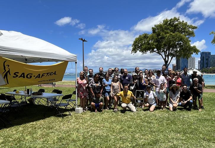 Un grupo grande y diverso de personas posa en un área de picnic cubierta de hierba con las aguas azules de la playa Ala Moana detrás de ellos. En primer plano hay un dosel con una pancarta amarilla que dice en letras negras “SAG-AFTRA”.