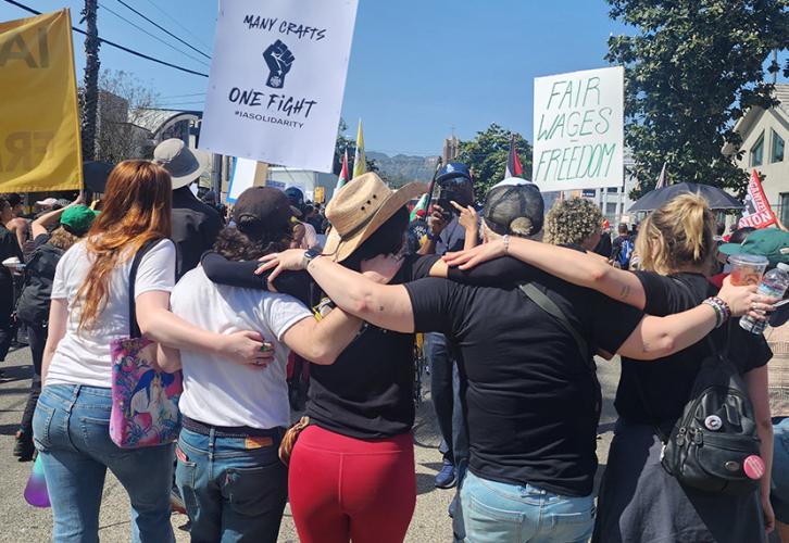 Five people, back to the viewer, with their arms around each other’s shoulders outdoors. Three individuals are in black T-shirts and two in white T-shirts. Union support signs can be seen in the background.