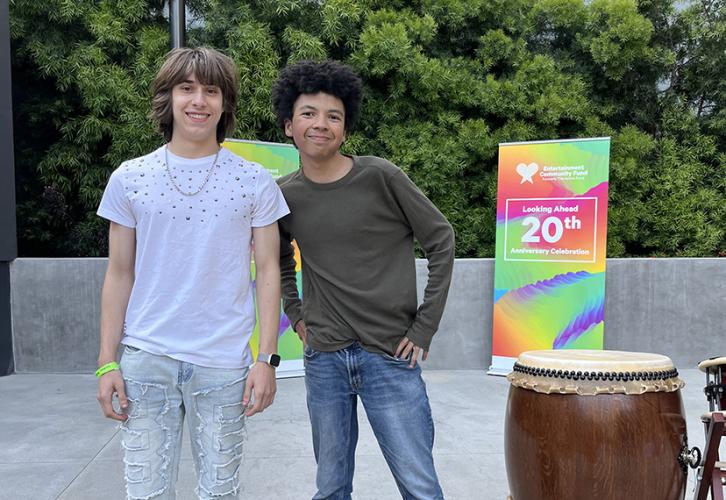 A white male wearing jeans and a white T-shirt and a mixed male wearing jeans and a olive green long sleeve T-shirt, standing next to a drum and a Looking Ahead 20th Anniversary/Entertainment Community Fund vertical banner.