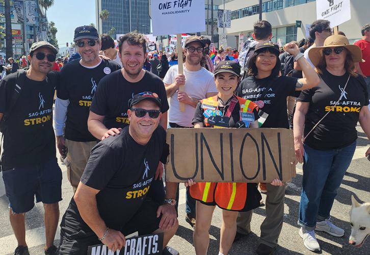 A large group of  people of different ethnicities pose outside with various union support signs. Many wear SAG-AFTRA or "SAG-AFTRA Strong" T-shirts.
