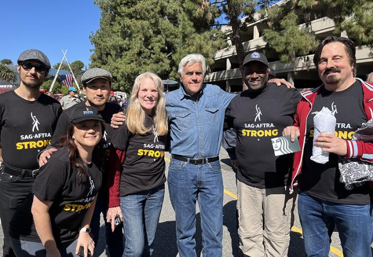 Men and women of different ethnicities wearing black "SAG-AFTRA Strong" shirts outside. Jay Leno, center, is dressed in a denim long sleeve shirt and jeans.
