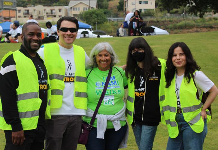 A group of five men and women of various ethnicities outside. A woman in the center is wearing a bright green shirt. The rest of the men and women around her are in black or white “SAG-AFTRA Strong” shirts with safety vests.